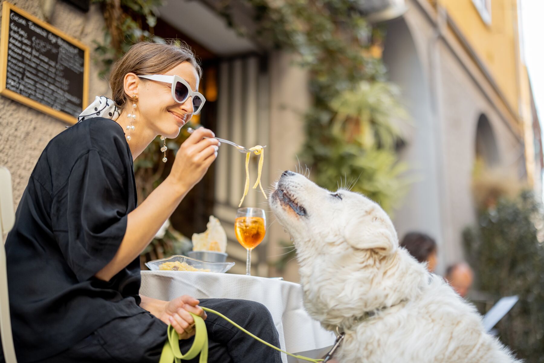 A woman sharing lunch with her dog at an outdoor table in Italy